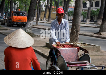 Ein Cyclo-Fahrer mit einer amerikanischen Baseballmütze fährt vorbei an eine Frau mit einem vietnamesischen Strohhut in Saigon, Vietnam. Stockfoto