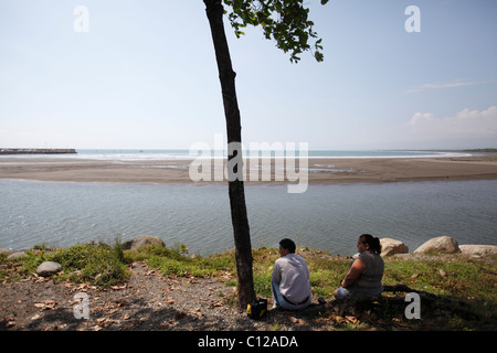 Ein Mann und eine Frau sitzen im Schatten eines Baumes mit Blick auf den Strand und den Eingang zum Hafen in Quepos, Costa Rica Stockfoto