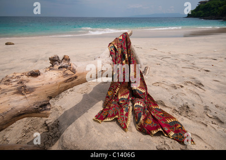 Eine Batik-Sarong drapiert über Treibholz auf dem weißen Sand Strand von Bias Tegal in Padang Bai auf Bali Stockfoto