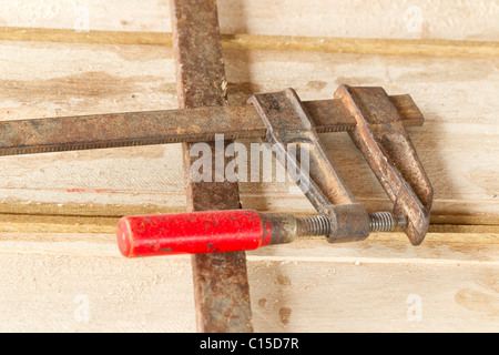 Nahaufnahme einer verwitterten, rostigen Metallklammer mit einem hellroten Griff auf einer hellen Holzoberfläche. Die Schelle verfügt über einen Schraubenmechanismus mit Gewinde und weist Anzeichen für starke Beanspruchung und auf Stockfoto