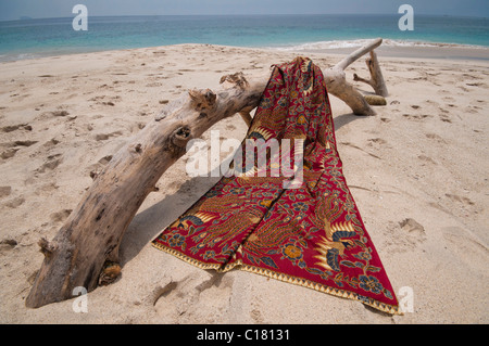 Eine Batik-Sarong drapiert über ein Treibholz-Protokoll auf dem weißen Sand Strand von Bias Tegal in Padang Bai, Bali Stockfoto