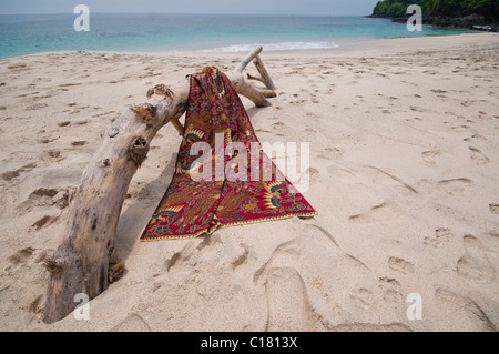 Eine Batik-Sarong drapiert über ein Treibholz-Protokoll auf dem weißen Sand Strand von Bias Tegal in Padang Bai, Bali Stockfoto