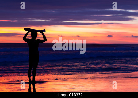 Eine Silhouette einer Person, die bei Sonnenuntergang an einem nassen Sandstrand steht und mit beiden Händen ein Surfbrett horizontal über dem Kopf hält. Der Himmel zeigt leuchtende Orange-, Rosa- und Violetöne Stockfoto