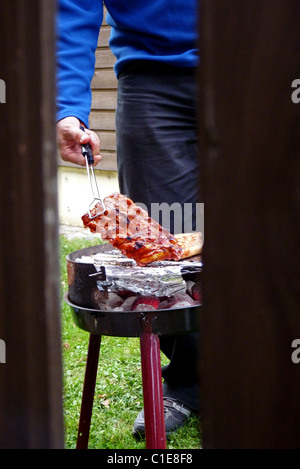 Mann, die Zubereitung von Fleisch auf dem Grill, gesehen durch einen hölzernen Zaun Stockfoto