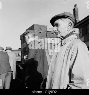 1950er Jahre, ein älterer Markthändler in Regenmantel und flacher Mütze, der draußen an Kisten auf Londons berühmtem Billingsgate Fish Market, Lower Thames Street, steht. Stockfoto