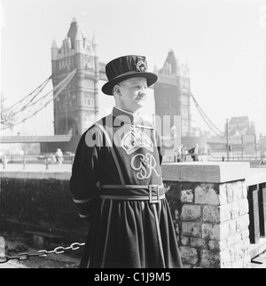 1950er Jahre, historisch, ein Tower of London Yeoman Warder oder beefeater, ein zeremonieller Leibwächter des englischen Herrschers mit Tower Bridge im Hintergrund. Stockfoto