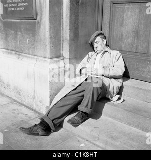 1950er Jahre, ein müder alter Mann, in Regenmantel und flacher Mütze, vielleicht ein Tramper, der nachmittags auf den Stufen des Hungerford House, Embankment, London schlummert. Stockfoto