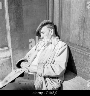 1950er Jahre, ein müder alter Mann, in Regenmantel und flacher Mütze, vielleicht ein Tramper, schlummert nachmittags auf den Stufen des Hungerford House, Embankment, London. Stockfoto