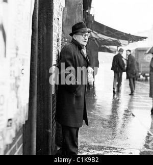 1950er Jahre London. Älterer Mann auf Gehweg in Camden Town, Hut und mit seinen Händen in seinen Mantel um ihn vor Regen zu schützen. Stockfoto