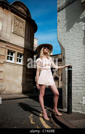 blonde Mädchen stehen in hellen sonnigen Sommer Straße trägt ein Seidenkleid und großen Hut mit Krempe und große Sonnenbrillen warten Stockfoto