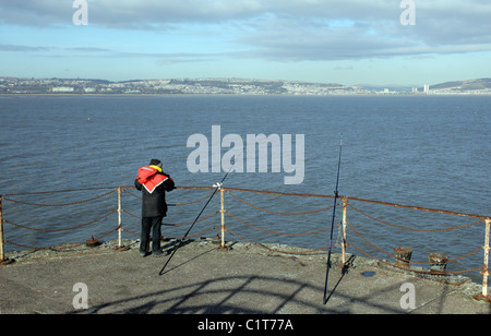 Ein Fischer mit seinem Angelruten ruhen neben ihm Ende weit Ende murmelt Pier mit Blick auf Swansea Bay, South Wales, Australia Stockfoto