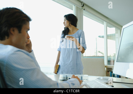Frau stehend mit Tasse Kaffee, Blick aus Fenster, Mann arbeitet auf Computer im Vordergrund Stockfoto