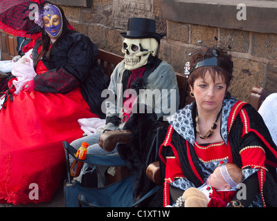 Zwei Frauen sitzen mit Baby-Puppen verkleidet als Goten und ein Mann in einer Schädel-Maske im Whitby Gothic Week End Frühling 2011 Stockfoto