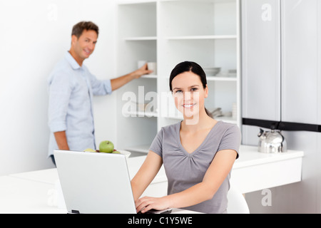 Glückliche Frau arbeitet mit einem Laptop, während ihr Ehemann Kaffeezubereitung Stockfoto