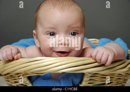 Niedliche Baby junge liegend auf Bauch in Wicker Wäschekorb. Er ist vier Monate alt und hat einen charmanten Ausdruck auf seinem Gesicht. Stockfoto