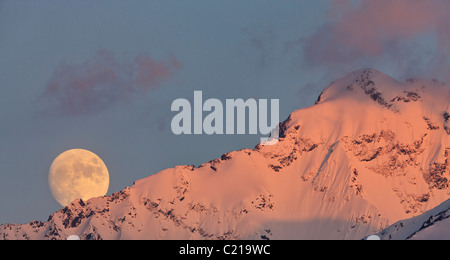 Ein fast Vollmond steigt hinter Eagle Peak im Alpenglühen vor Sonnenuntergang in Mitte des Frühlings in den Chugach Mountains von Alaska. Stockfoto