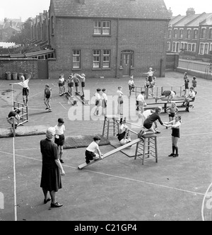 Im Jahr 1950s überwacht eine Lehrerin ein kleines Kind, das eine Sportausbildung außerhalb eines innerstädtischen Grundschulspielplatzes in England macht. Stockfoto