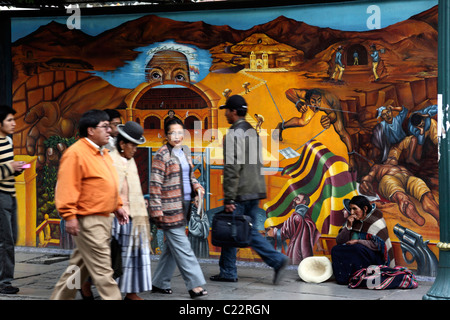 Menschen, die an einer Dame aus der Region Potosi vorbeigehen und vor einem Wandgemälde betteln, das die Repression von Ureinwohnern in den Silberminen von Potosi in der Vergangenheit, La Paz, Bolivien, zeigt Stockfoto