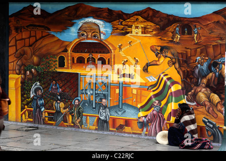 Dame aus der Region Potosi bettelte vor einem Wandgemälde, das die Repression von Eingeborenen in Potosi Silberminen in der Kolonialzeit, La Paz, Bolivien zeigt Stockfoto