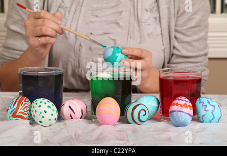 Eine junge Frau malt sorgfältig ein buntes Osterei hinter vielen anderen und drei Gläser von farbigen Farbstoff. Stockfoto