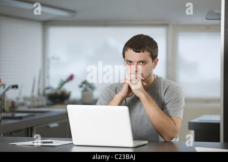 Junger Mann sitzt stützte sich auf Ellenbogen am Laptop in Küche Stockfoto