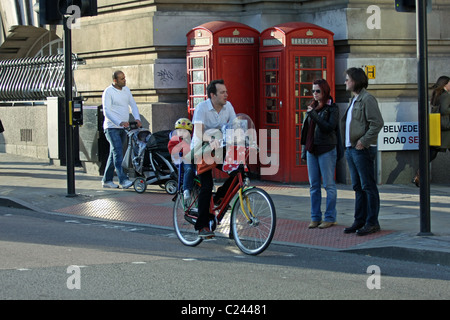 Ein Mann, mit dem Fahrrad, mit einem Kind in einem Sitz hinter ihm, Menschen zwei warten darauf, überqueren Sie die Straße, ein Mann, ein Kinderwagen schieben Stockfoto