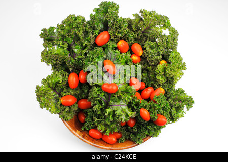 Kleine rote Kirschtomaten auf roten Grünkohl in einer hölzernen Schüssel auf weißem Hintergrund. Helle und kleine rote Tomaten und grünem Kohl platziert in einer Holzschale Stockfoto