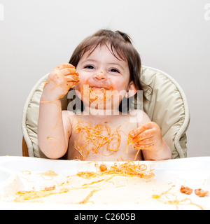 Glückliches lächelndes Baby Spaß Essen chaotisch bedeckt in Angel Hair Pasta Spaghetti rot marinara Tomatensauce. Stockfoto