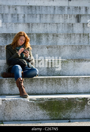 Junge Frau setzte sich auf konkrete Schritte mit einem Mobiltelefon. Stockfoto