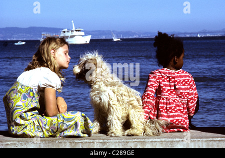 Zwei kleine Mädchen, gekleidet in Retro 60er Jahre Kleidung sitzen auf San Francisco Bay Pier mit zottigen Hund spielen Stockfoto