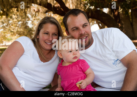 Mutter und Vater mit acht Monate alten Tochter posieren für die Kamera im Schatten eines Baumes Stockfoto