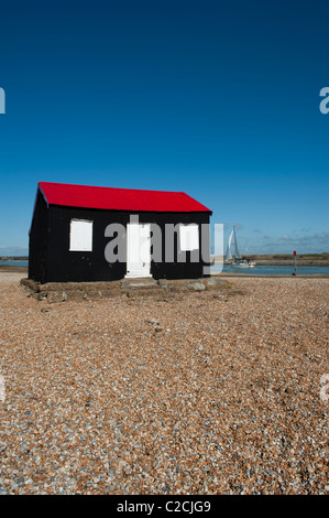 Eine weiße Yacht segelt vorbei eine rote und schwarze gewellte Hütte mit weißen Türen auf der Kiesstrand am Roggen Hafen gebaut. East Sussex Stockfoto