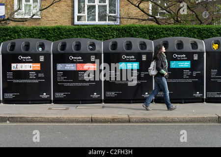 Junge Frau, die zu Fuß durch eine Reihe von Recycling-Behälter, London, England, UK Stockfoto