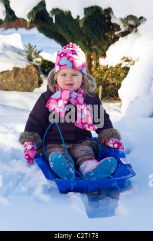 Vertikale Porträt eines Mädchens gewickelt Warm mit passendem Schal, Mütze und Handschuhe spielen auf einer Rodel oder Schlitten im Schnee Stockfoto