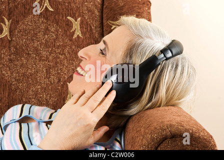 Alte Frau mit Kopfhörern Musik hören und lachen, She auf Sofa im Wohnzimmer entspannen Stockfoto