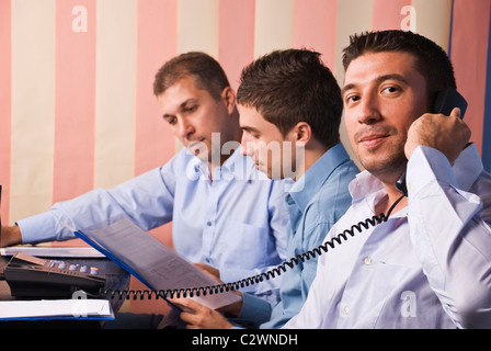 Drei Unternehmer in Büro, konzentrieren sich auf erste Mensch, der Telefon, die beiden anderen Männer lesen und haben eine Gespräch spricht Stockfoto