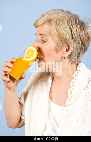 Gesunde ältere Frau im Profil stehen und trinkt ein Glas mit frisch gepresstem Orangensaft auf blauem Hintergrund Stockfoto