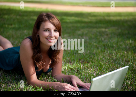Lächelnde junge Frau auf Bauch liegend funktioniert auf Computer Laptop auf dem grünen Rasen ihres Hauses. Stockfoto