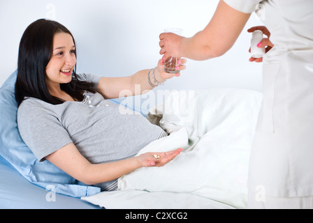 Schwangere Frau liegend auf dem Bett im Krankenzimmer und eine Krankenschwester, die ihre Vitamine und ein Glas mit Wasser geben Stockfoto