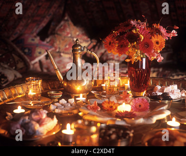 Marokkanischen Stil Messing Tee-Set auf Tisch mit Kerzen auf Messing Tischplatte und rosa und roten Blumen in Glasvase. Stockfoto