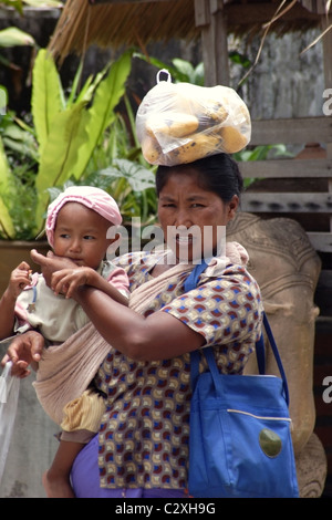 Eine Frau trägt Früchte oben auf dem Kopf und ein Baby in ihren Armen in Nordthailand. Stockfoto