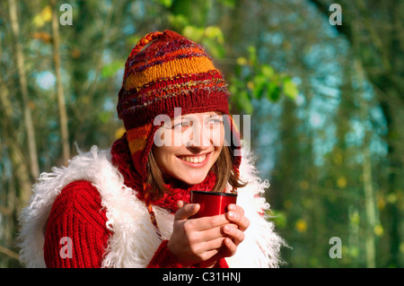 JUNGE FRAU, DIE SICH AUFWÄRMEN MIT EINEM HEIßEN GETRÄNK WÄHREND AUF EINEM SPAZIERGANG IM WALD Stockfoto