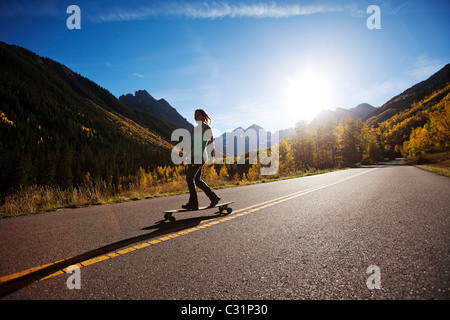 Eine junge Frau Longboards auf einer glatten Landstraße durch die Berge und die goldenen Wälder. Stockfoto