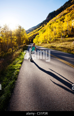 Eine junge Frau Longboards auf einer glatten Landstraße durch die Berge und die goldenen Wälder. Stockfoto