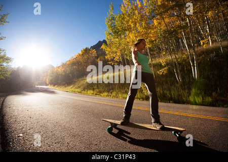 Eine junge Frau Longboards auf einer glatten Landstraße durch die Berge und die goldenen Wälder. Stockfoto