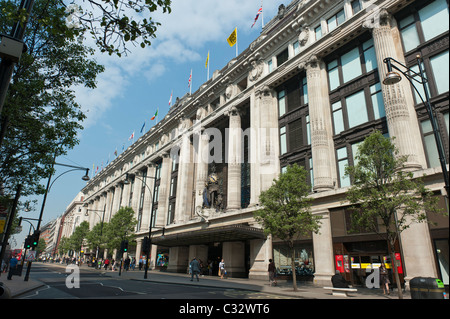 Shopper gehen auf Oxford Straße, vorbei an der Queen of Time Skulptur über dem Art-deco-Eingang in Selfridges laden. Stockfoto