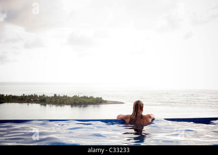 Eine Frau sitzt mit ihren Armen auf den Rand des einen Infinity-Pool mit Blick auf den weiten Ozean. Stockfoto