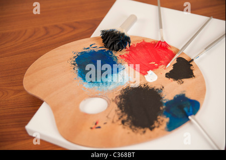 Eine Holzpalette mit gemischten Farben Farben und Pinsel sitzt oben auf einer leeren Leinwand in einem Kunstatelier. Stockfoto