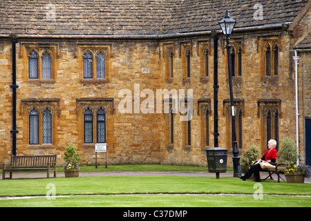 Die reife Frau saß auf der Bank vor dem Stein Gebäude in Sherborne im April - Almshüuser aus dem 15. Jahrhundert Behütete Unterkünfte für ältere Menschen Stockfoto