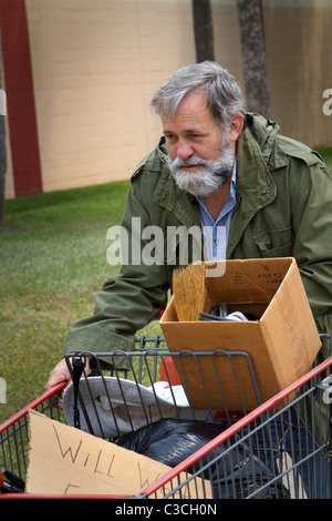 Obdachloser Mann trägt einen alte Armeemantel schiebt einen Einkaufswagen hält seinen Besitz. Stockfoto
